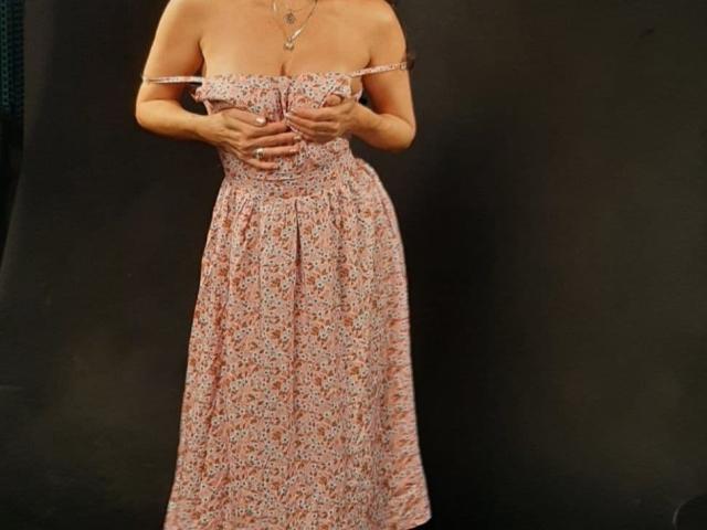Les mains d'Antralyn couvrent ses seins, elle porte une robe fleurie rose avec des bretelles qui tombent d'une épaule, et elle se tient devant un fond sombre. Elle porte un collier délicat..