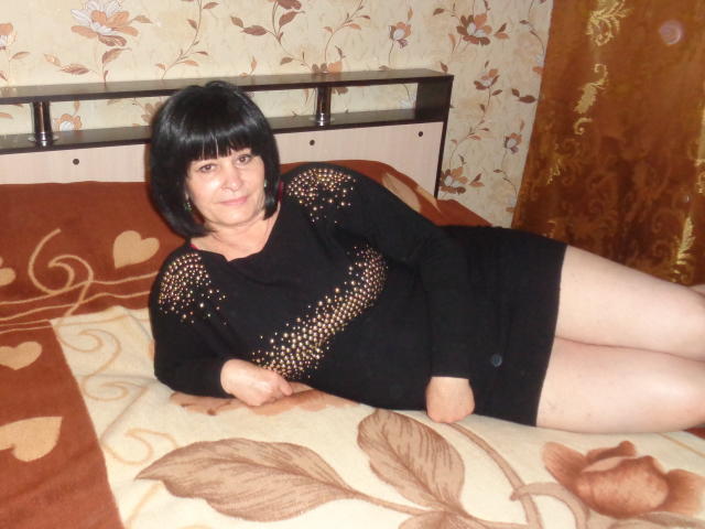 AhVredinka, avec sa coupe de cheveux noire, portant une robe noire avec des motifs troués, est allongée sur un lit à motifs marron et beige, montrant ses jambes pâles et un petit bouton noir..