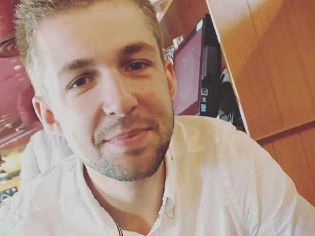 HomTeo, com cabelo castanho curto, barba clara e camisa branca, sorrindo levemente, mostrando pele clara e olhos castanhos, em um selfie casual..