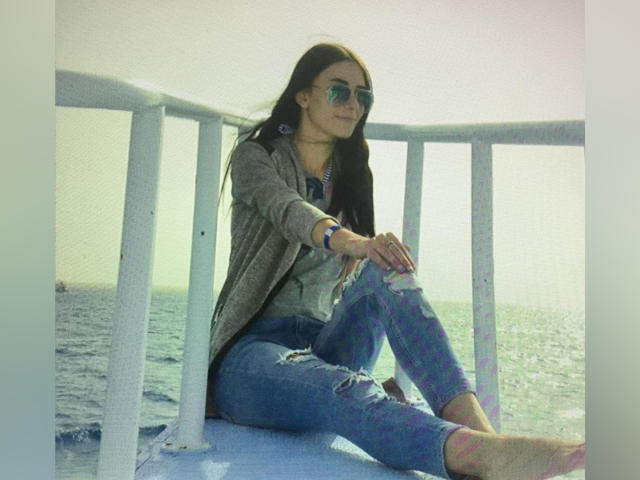 Nika, con lunghi capelli scuri, indossa occhiali da sole, un cardigan grigio, jeans blu strappati e un braccialetto blu, seduta su una ringhiera bianca, mentre guarda verso la fotocamera..