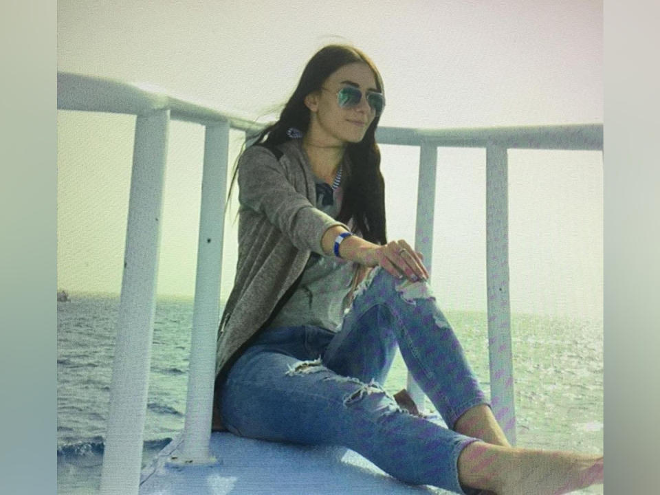 Nika, avec de longs cheveux noirs, portant des lunettes de soleil, un cardigan gris, un jean bleu déchiré et un bracelet bleu, assise sur une rambarde blanche, regardant l'appareil photo..