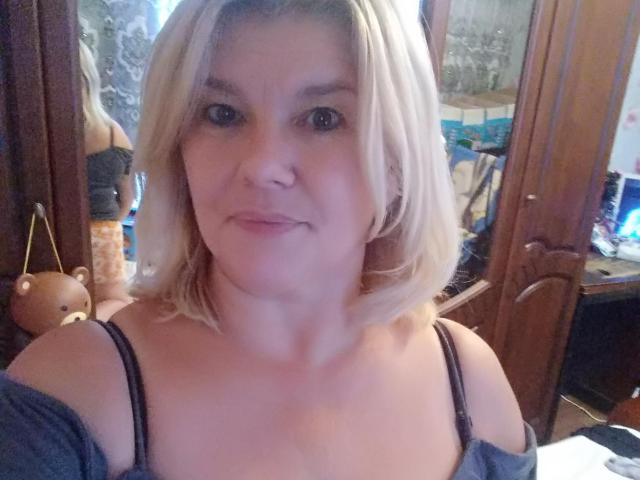 DreamyShirley, con cabello rubio corto, piel clara, ojos azules y pequeños senos, vestida con un top negro de tirantes, sonriendo suavemente, en una habitación de madera con un espejo y un osito de peluche..