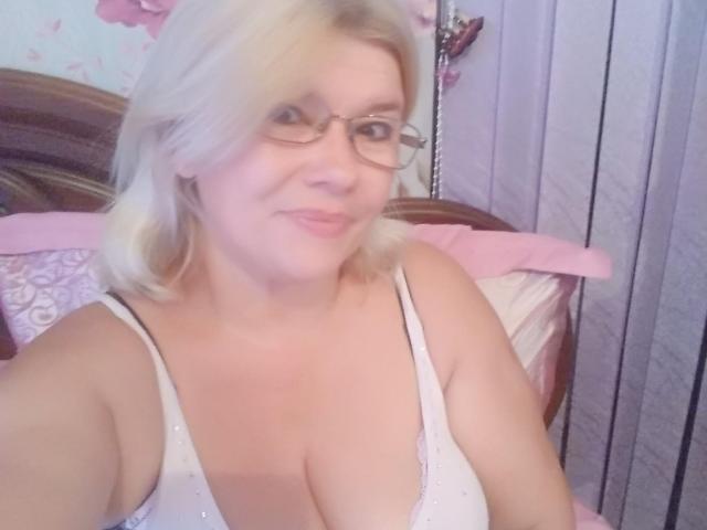 DreamyShirley, com cabelo loiro, óculos e uma camiseta branca que mostra seus seios grandes, redondos e nus. Ela está sorrindo suavemente, olhando para a câmera..