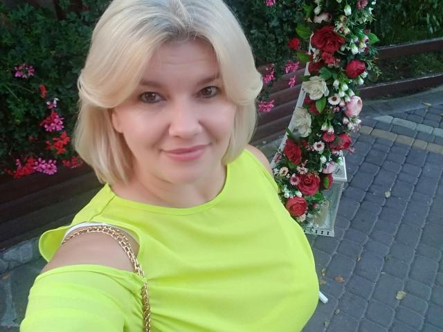 DreamyShirley, con i capelli biondi, indossa una maglietta giallo neon, sorride, tiene una catena d'oro, mostra una spalla scoperta e si trova vicino a una colonna decorata con fiori. Il seno non è visibile..