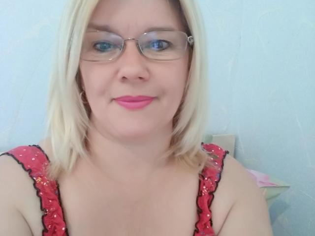DreamyShirley con cabello rubio, gafas, lápiz labial rosa y una blusa roja con lentejuelas, mostrando sus senos, sonriendo ligeramente, ojos azules, piel pálida, en un selfie..