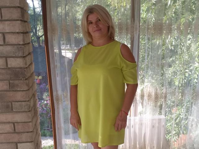 DreamyShirley stands in a bright yellow dress with cut-out shoulders, looking slightly annoyed. She has blonde hair and a medium build. Her tits are small, and she's bare-assed.