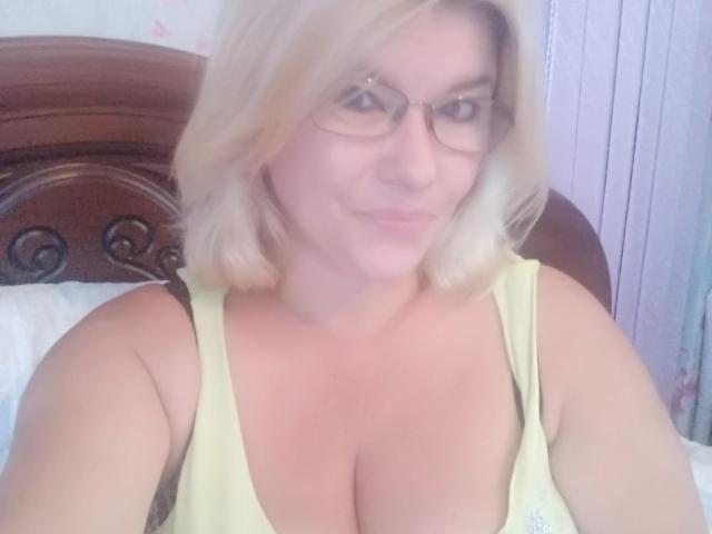 DreamyShirley, com cabelo loiro, óculos e uma regata amarela que mostra seus seios grandes e claros. Ela está sorrindo levemente, com um olhar sutil e sexy..