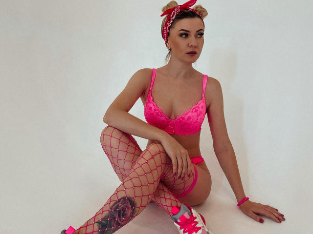 ParadiseRoxanne mit rosa BH, rosa Netzstrümpfen, rosa Schleifenhaarreif, rosa Armband, sitzend, die Brüste zeigen, kleines Tattoo an ihrem rechten Oberschenkel, zur Seite blickend..
