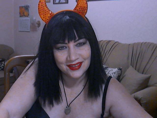 MelissaLight mit schwarzem Bob, orangefarbenen Teufelshörnern, rotem Lippenstift, schwarzem Tanktop, Halskette, lächelnd in die Kamera, blasse Haut, mittelgroße Brüste, leicht mollig, Wohnzimmer, ungezwungene Umgebung..