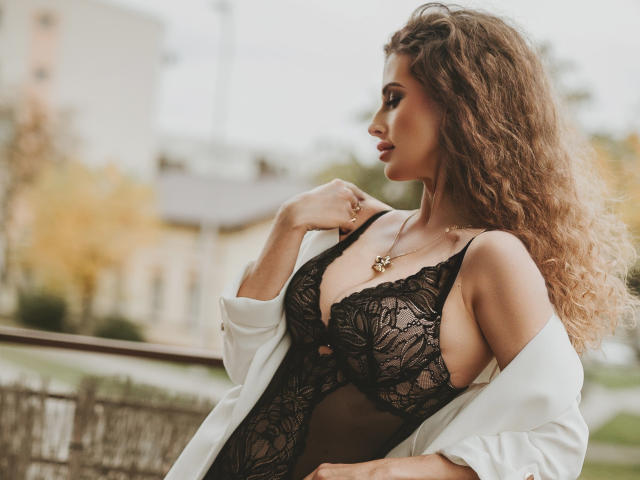MikaFetish met krullend bruin haar, gekleed in een zwart kanten body, witte blouse open, borsten zichtbaar, gouden ketting, profiel van de zijkant, hand op het sleutelbeen, buiten, overdag, sexy..