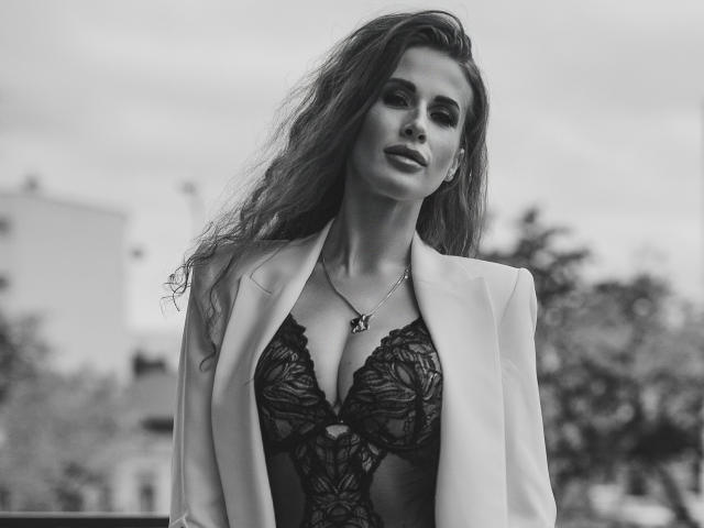 Black-and-white photo of MikaFetish with long, wavy hair, wearing a lace bodysuit and open blazer, showing her tits and a necklace with a small pendant.