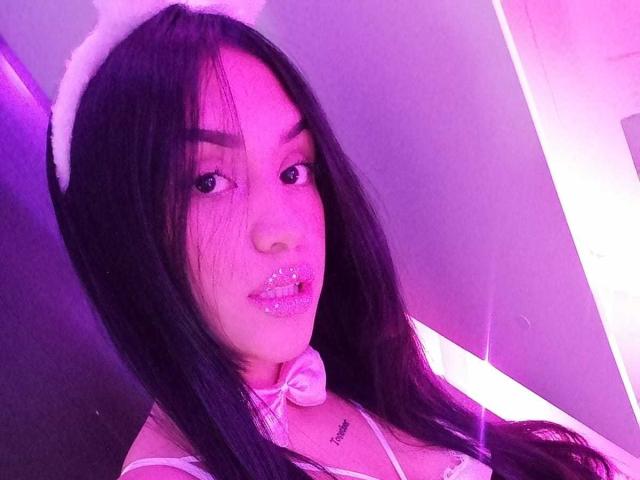 Samantha Sweett, con cabello negro largo, lleva una diadema con orejas de gato blancas y brillo labial brillante, un collar con lazo blanco y un tatuaje debajo del mentón. Iluminación rosa..