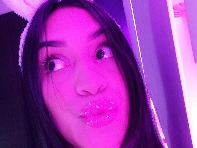 Il viso di SamanthaSweett, con il trucco illuminato di viola, i capelli neri e le labbra rosa scintillanti arricciate, indossa una fascia con orecchie da coniglio bianche, guardando leggermente di lato con occhi spalancati..