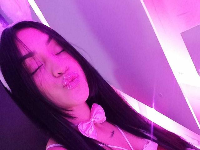 Samantha Sweett mit langem, schwarzem Haar, geschlossenen Augen, vollen, glitzernden, rosafarbenen Lippen, einer weißen Schleife und einem kleinen Tattoo auf der Brust, beleuchtet von violettem Neonlicht..
