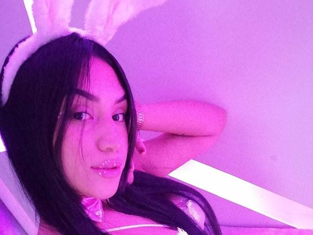 SamanthaSweett, con i capelli neri, indossa orecchie da coniglio bianche, rossetto rosa glitterato e una collana rosa a fiocco, posa con un braccio dietro la testa, sotto luci viola..