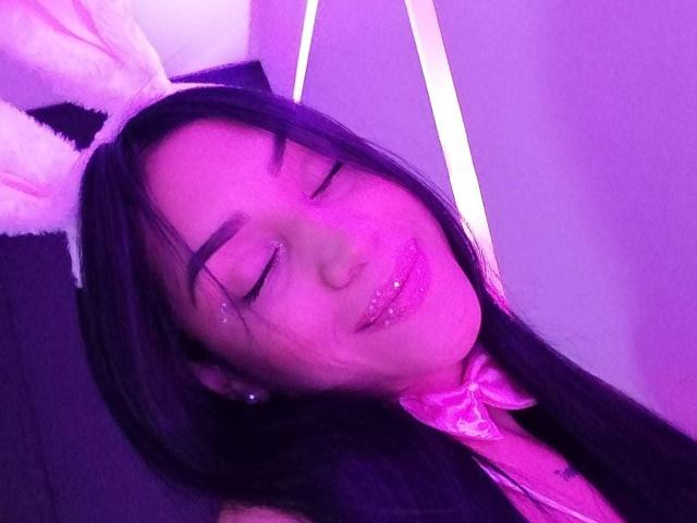 SamanthaSweett, con il viso leggermente violaceo, gli occhi chiusi e un sorriso carino, indossa un fiocco rosa e orecchie da coniglio bianche, ha delle paillettes sulla guancia e lunghi capelli neri..