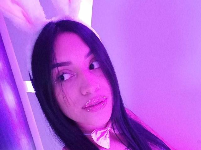 SamanthaSweett, avec des cheveux noirs, une lumière violette scintillante, portant des oreilles de lapin blanches et duveteuses, des lèvres roses brillantes et un nœud papillon rose, regardant sur le côté, avec une expression mignonne et séduisante..