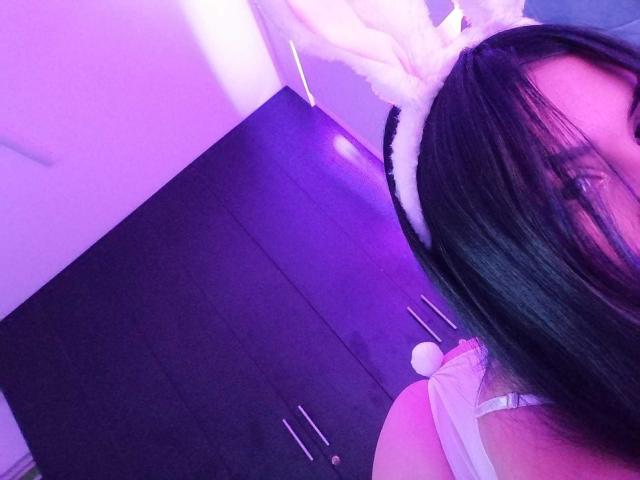 SamanthaSweett, com cabelo preto, usando orelhas de coelho brancas e uma blusa rosa, mostrando a parte de trás branca de um coelho, com iluminação roxa. Seios parcialmente visíveis..