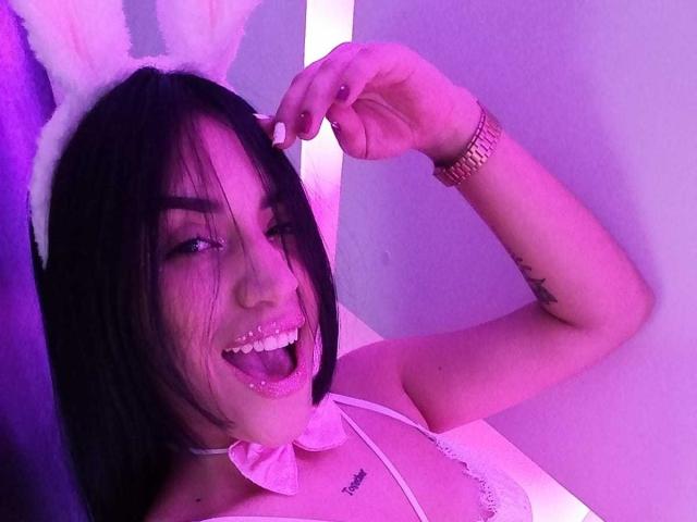 SamanthaSweett, com cabelo preto, glitter rosa na língua, usando orelhas de coelho brancas, um laço rosa e uma blusa branca, sorrindo e tocando sua orelha com a pele que brilha em rosa..