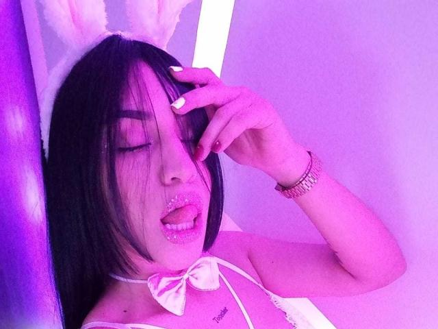 SamanthaSweett, com cabelo preto, orelhas de coelho rosa, laço branco e lábios brilhantes, tocando o rosto, com iluminação roxa, exibindo suas unhas brancas e brilhantes e batom rosa..