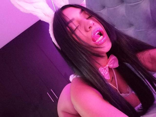 SamanthaSweett, avec ses longs cheveux noirs, ses lèvres pailletées roses, ses oreilles de lapin blanches, son nœud papillon rose et sa lingerie blanche, exhibant sa poitrine et sa bouche ouverte, dans une pièce éclairée en violet..