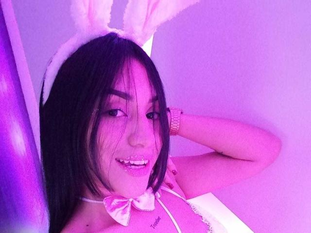SamanthaSweett, com cabelo preto, orelhas de coelho brancas, dentes brilhantes rosa e um laço rosa, vestindo uma blusa branca, olhando para a câmera com uma mão atrás da cabeça..
