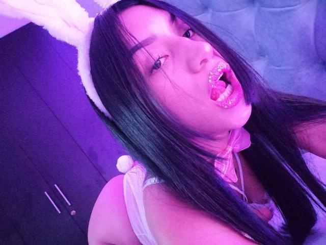 SamanthaSweett, com cabelos pretos longos, orelhas de coelho brancas e um colar rosa, língua de fora, mostrando dentes brilhantes, vestindo lingerie branca, com um plug anal de silicone em formato de rabo fofo. Selfie com iluminação roxa..
