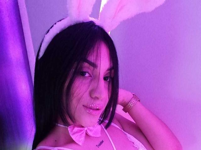 SamanthaSweett, avec des cheveux noirs, des oreilles de lapin blanches, un rouge à lèvres pailleté rose, un haut en dentelle blanche, un nœud rose et une montre dorée, regardant de manière séduisante l'appareil photo, dans un éclairage violet..