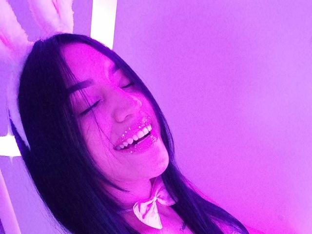 SamanthaSweett, con i capelli neri, glitter rosa sulle labbra, orecchie da coniglio e un fiocco bianco, sorride con gli occhi chiusi, illuminata da luci viola. Il suo seno è leggermente visibile..