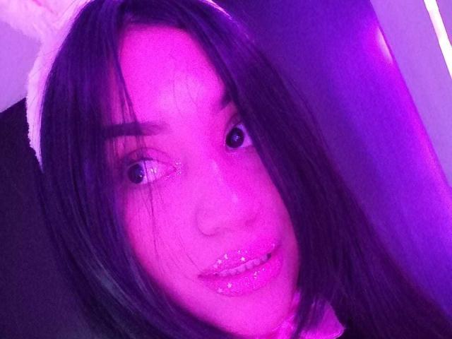 SamanthaSweett, con il viso illuminato di viola, lunghi capelli neri, orecchie da coniglio bianche, labbra rosa scintillanti e occhi lucidi, sorride leggermente. Il seno non è visibile..