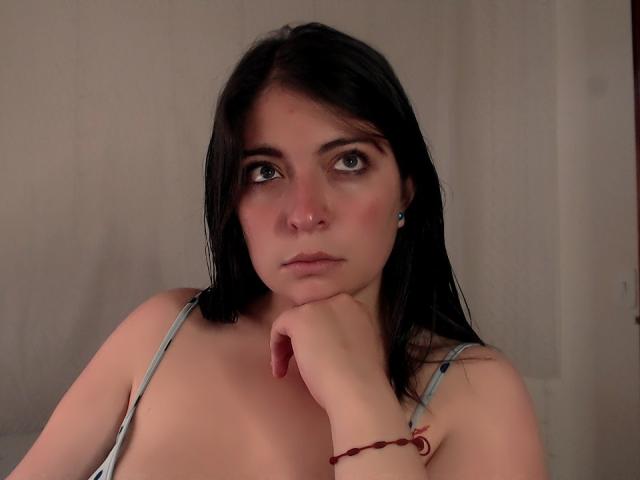 BrightLightz, con capelli scuri, occhi azzurri, piercing al naso e braccialetto rosso, appoggia il mento sulla mano, indossa una maglietta grigia, mostrando il petto scoperto e piccoli seni, guardando leggermente di lato..