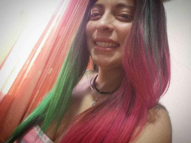 BrightLightz, con cabello largo rosa y verde, sonriendo, mostrando los dientes, vistiendo una blusa a rayas rosa y blanca, un collar tipo "choker" y con un ligero bronceado, en un selfie..