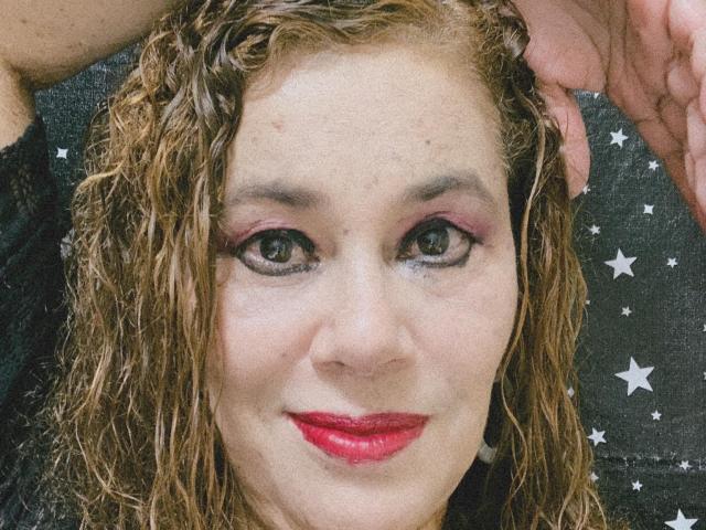 MadelaineLu has wet, curly brown hair, pink eye makeup, and red lipstick. Her hand is in her hair, showing off her green eyes and slightly smiling face.