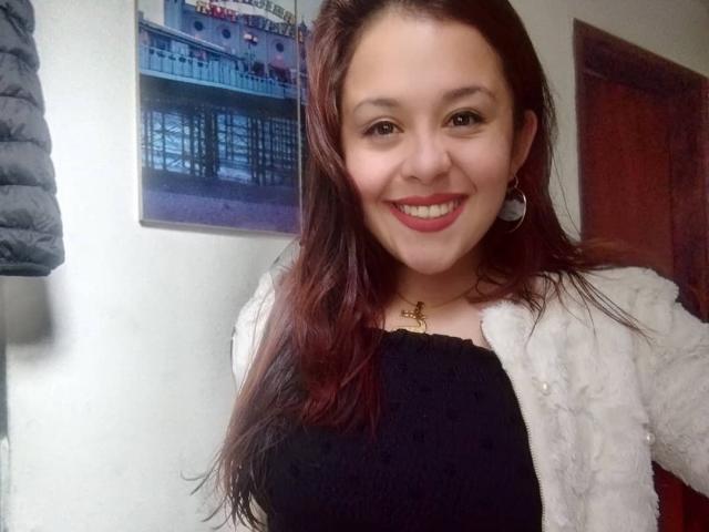 Katie Woolf, con cabello rojo, sonriendo, vestida con una blusa negra y una chaqueta blanca y suave, con aretes, collar, pechos pequeños y lápiz labial rojo, en un selfie..