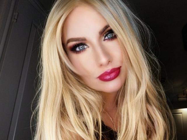 TinaBella has blonde hair, blue eyes, dark eyeliner, and bright pink lipstick. She's looking at the camera with a slight smile. Her tits are not visible.