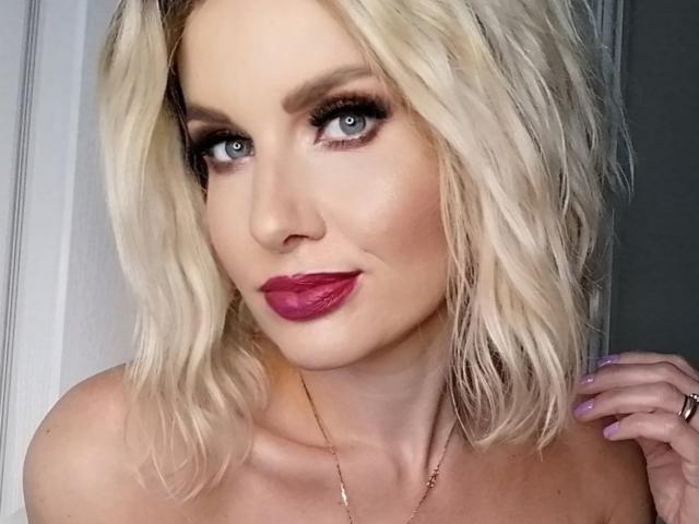 TinaBella with blonde wavy hair, blue eyes, and pink lipstick, smiling at the camera, showing off her tits and a gold necklace, with a subtle ring on her finger.