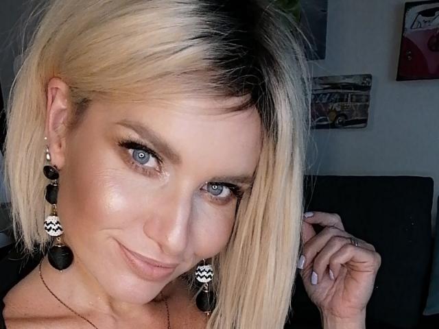 TinaBella has blonde hair with black highlights, blue eyes, and black zigzag earrings. She's smiling with a slight smirk, wearing a black top, and touching her hair.