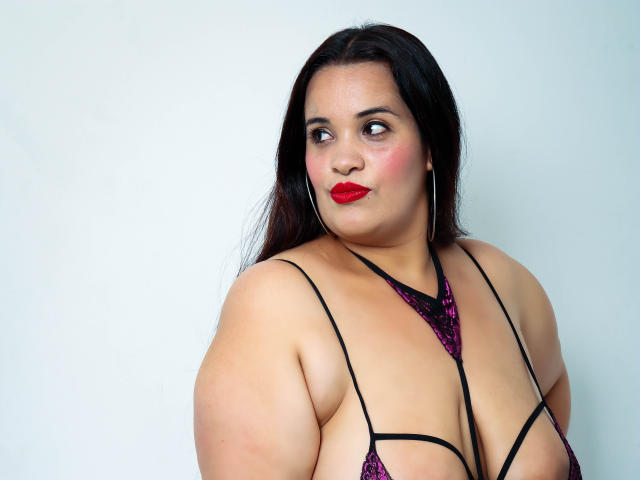 AloondraZaenss, de talla grande, con cabello oscuro, lápiz labial rojo y un sujetador de encaje púrpura, mirando hacia un lado, mostrando sus grandes senos y sus fuertes brazos sobre un fondo liso..