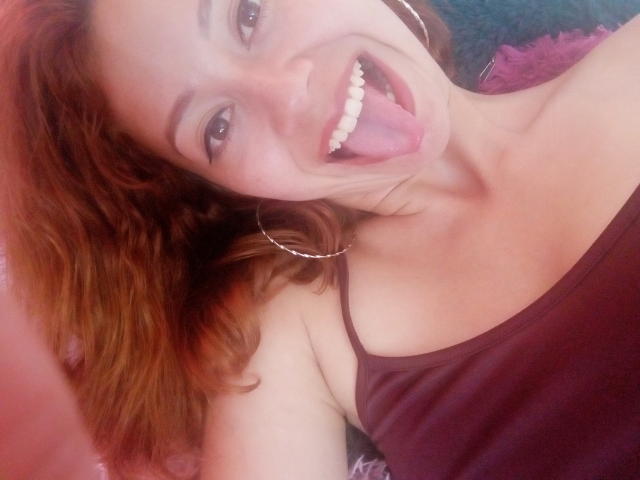 ElizaJones with red curly hair, sticking out her tongue, smiling, wearing a purple tank top and hoop earrings, showing her white teeth. Her boobs are partially visible.