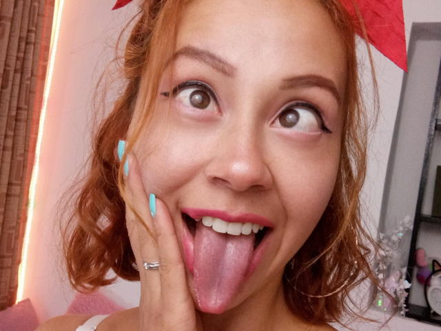 Eliza Jones, com cabelo ruivo, unhas azuis e uma faixa de cabelo vermelha, com a língua de fora, olhos arregalados e uma pequena linha preta abaixo do olho direito. Ela está tocando sua bochecha..