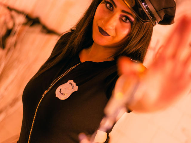 Charlotte Baker, dunkler Lippenstift, schwarzes Hemd mit Reißverschluss, Polizeihut, langes braunes Haar, hält einen Schlagstock, lächelt, mittelgroße Brüste, weiches orangefarbenes Licht, unscharfer Vordergrund, sexy, selbstbewusst, leicht verschmitzt..