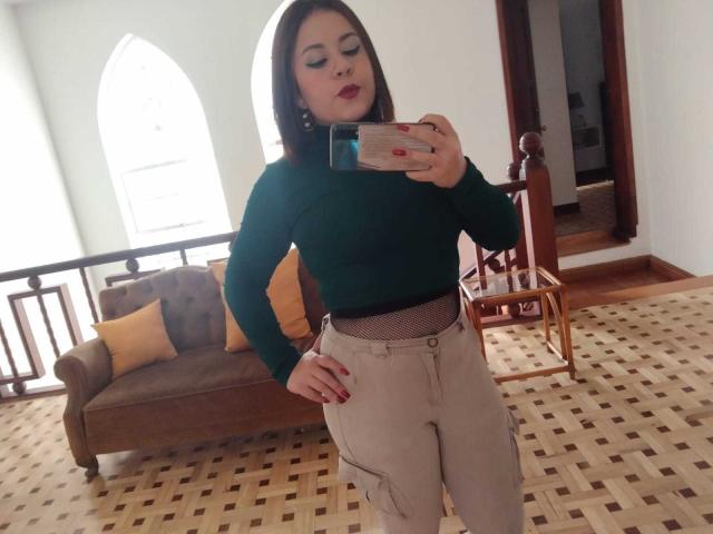 Katie Woolf, con i capelli castani, un maglione a collo alto verde e pantaloni beige, sta scattando un selfie allo specchio, con una mano appoggiata al fianco, unghie rosse, in una stanza con un divano marrone e un pavimento a motivi..
