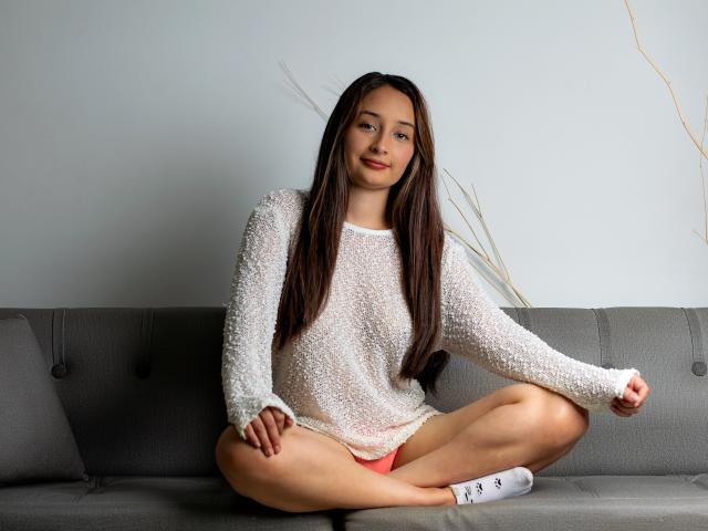 BeckyMils, zittend op een grijze bank, gekleed in een witte, gestructureerde, lange mouwen trui, met roze onderbroek zichtbaar, met lang bruin haar en witte sokken, glimlacht zachtjes..