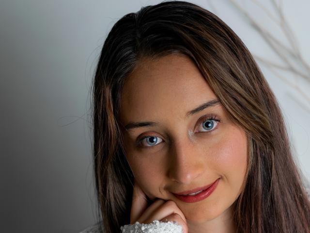 BeckyMils mit blauen Augen, braunen Haaren und einem süßen Lächeln, die ihr Kinn in die Hand legt, wobei sie ihre vollen, roten Lippen und ihre glatte, mit Sommersprossen übersäte Haut zur Schau stellt..
