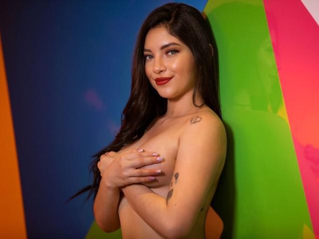 LaurenMaxwel, avec ses longs cheveux noirs, son rouge à lèvres rouge et ses tatouages, cachant ses seins avec ses mains, souriant à la caméra, debout devant un fond coloré..