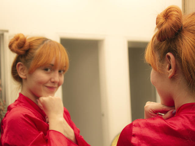 MollyMarmalade with red hair in buns, wearing a red shirt, smiling at her reflection in a mirror, hand under chin, soft light, casual, cute, slight blush.