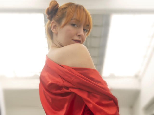 MollyMarmalade with red hair in a bun, wearing a red off-shoulder top, looking over her shoulder, showing her bare shoulder and a slight smile. Soft, natural light.