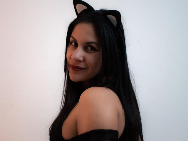 RoraimAnaggie mit schwarzen Katzenohren, langen schwarzen Haaren und rotem Lippenstift, lächelt in die Kamera und zeigt ihre nackten Schultern und ein schwarzes Oberteil, wobei ihre Brüste leicht sichtbar sind..