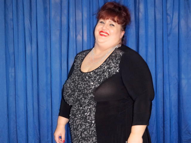 A plus-sized MilfClau with red hair, wearing a black and silver sequined dress, smiling with red lipstick, showing her large tits through sheer fabric. Blue curtain background.