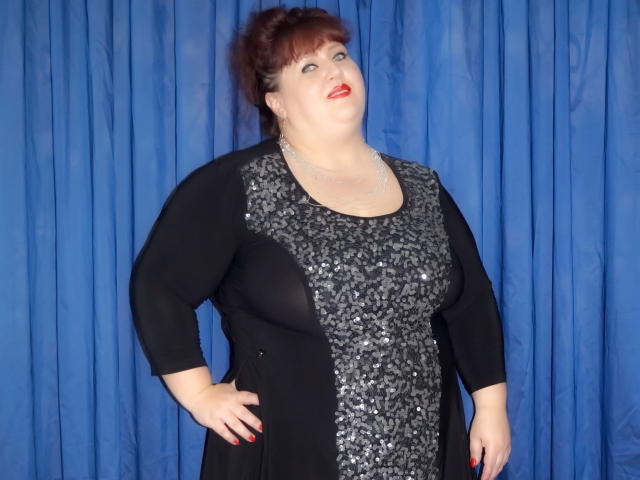A plus-sized MilfClau with red lipstick, wearing a black dress with silver sequins, standing against a blue curtain, has her hand on her hip, smiling.